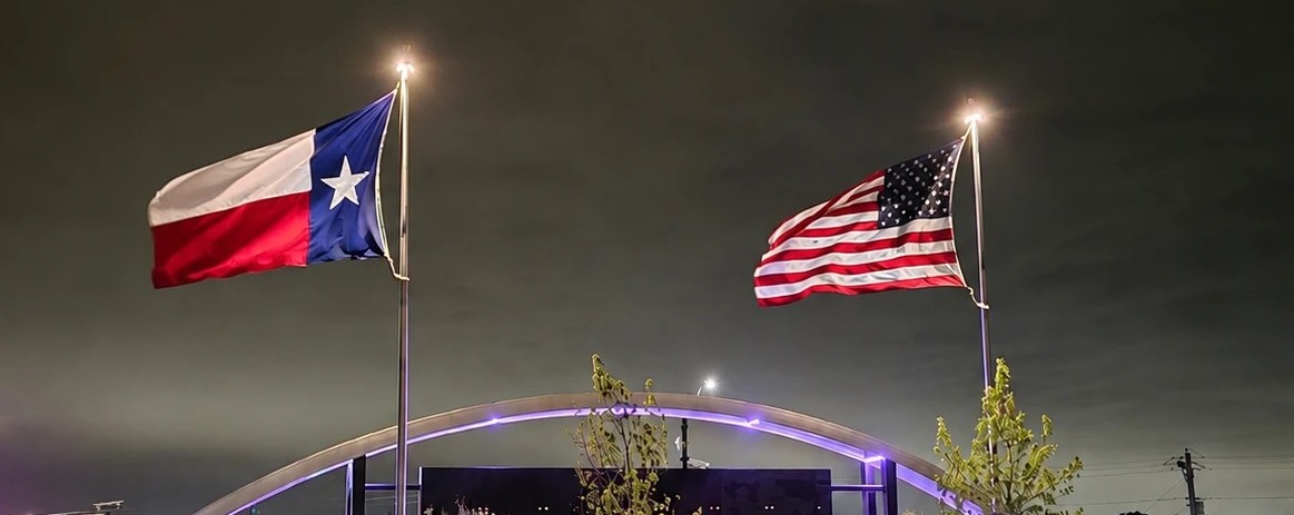 Two FlagCo Beacon flagpole lights illuminating the US and Texas flags after dark at a commercial entrance